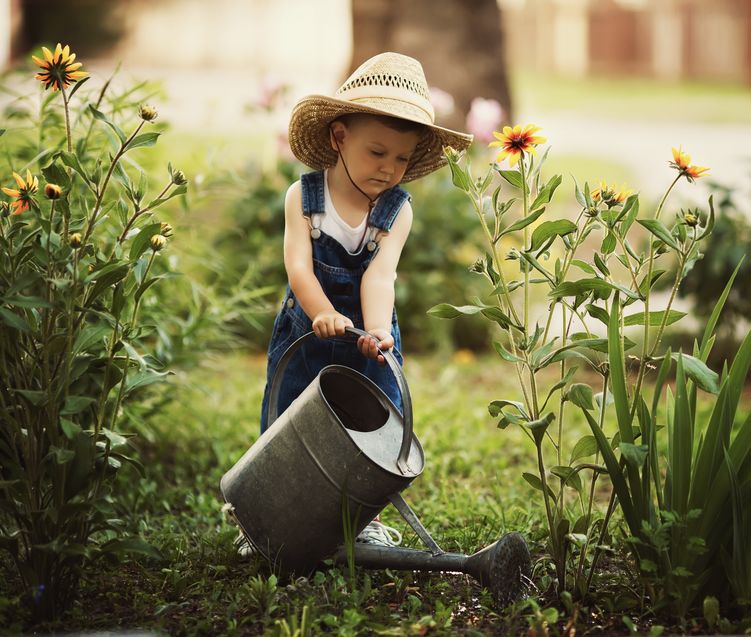 little boy wearing a floppy hat watering flowers with a huge watering can watering can