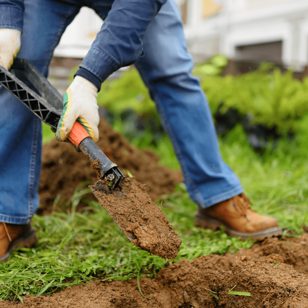 Mans' legs and hand holding a shovel over an open hole in the soil.